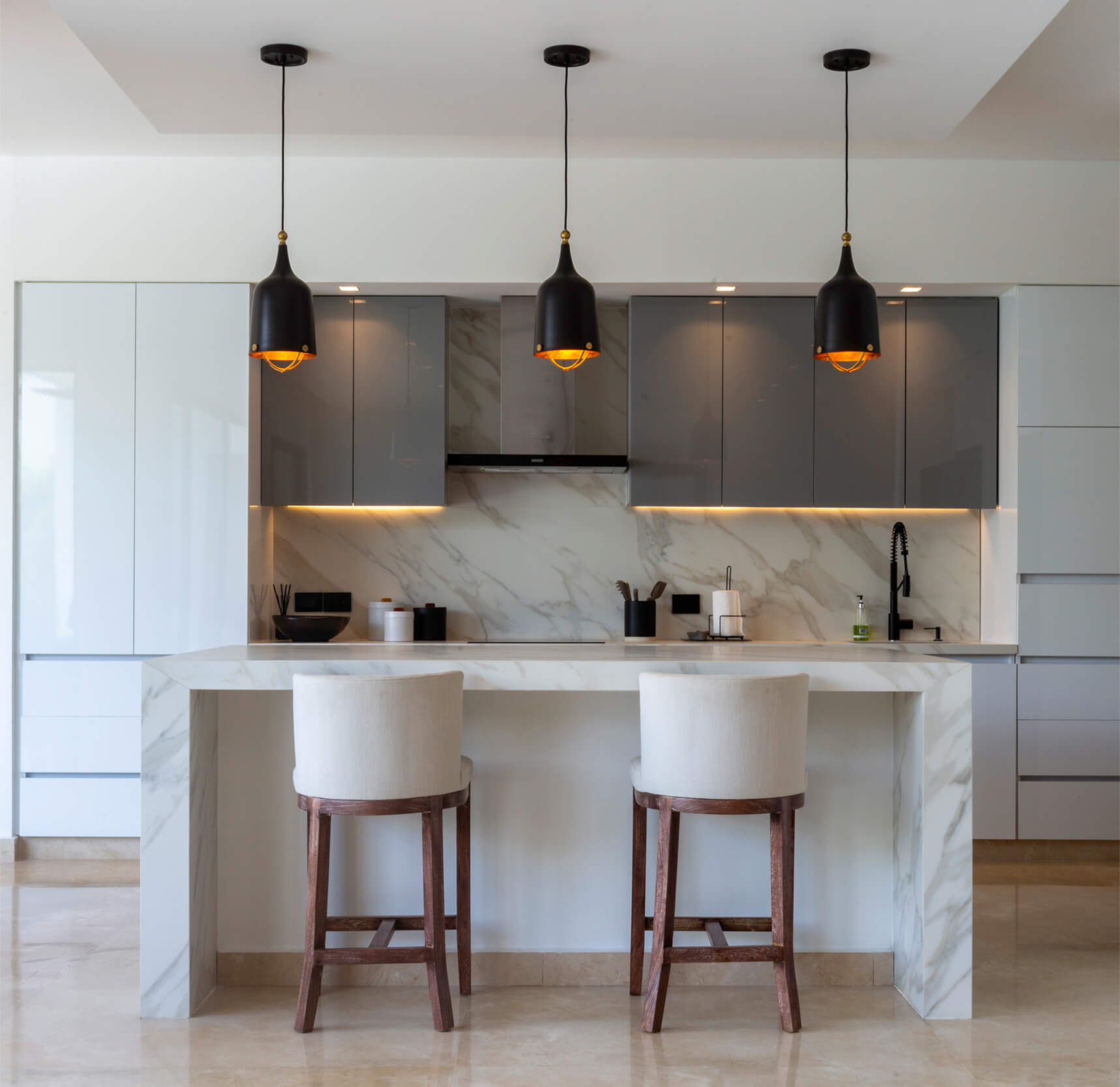 Cocina abierta de Casa Sarria 15|04 con isla central tipo mármol, muebles blancos, gabinetes grises y lámparas colgantes negras. Arquitectura residencial moderna.