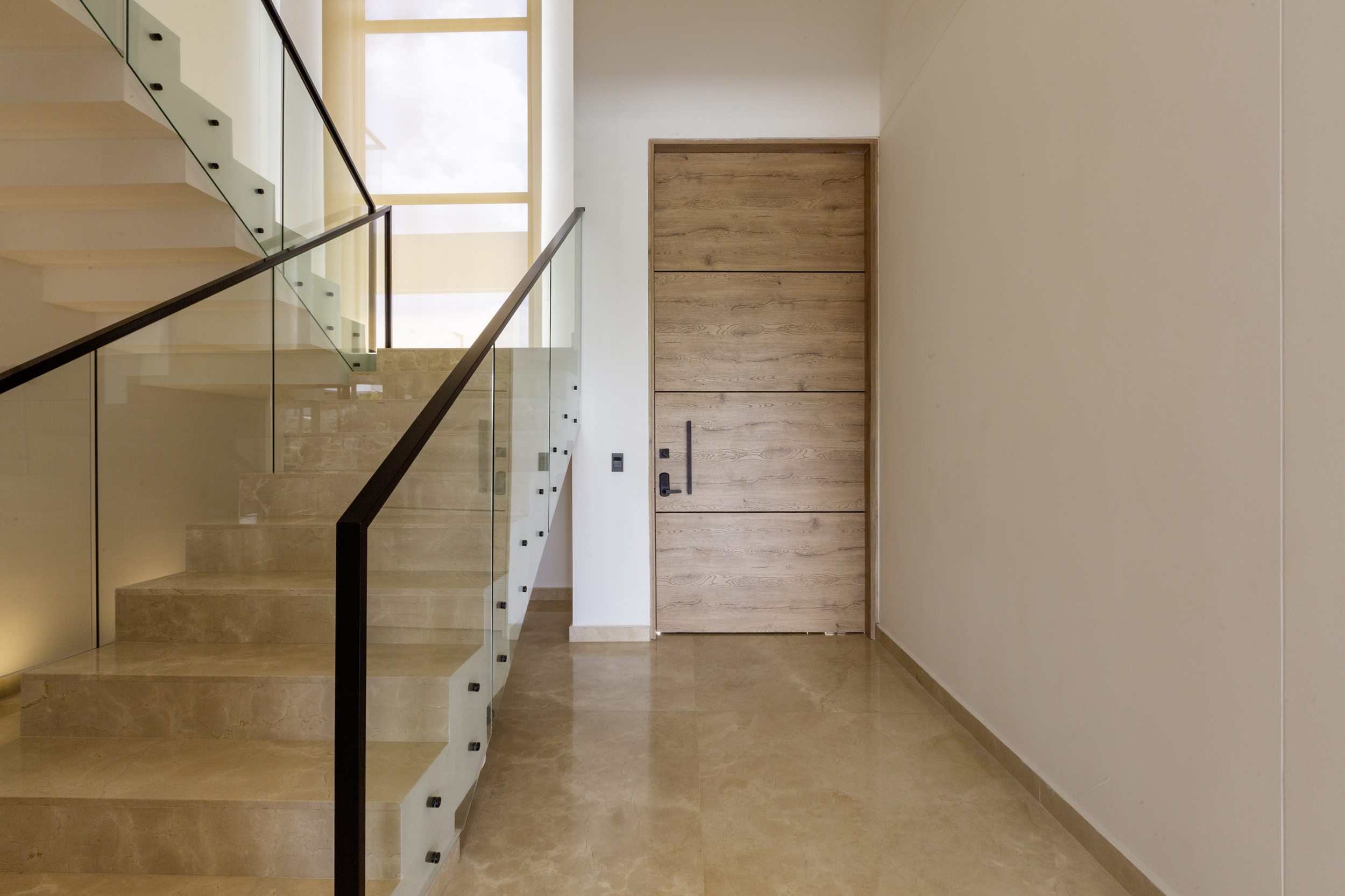Entrada principal de Casa Sarria 15|04 con escalera en mármol, baranda de vidrio, pasamanos negro y puerta en madera de diseño contemporáneo.